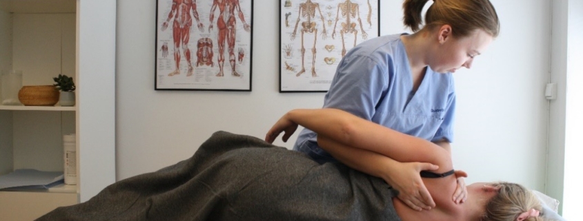 Bilde fra klinikk hvor osteopat Elisabeth behandler en kvinnelig pasienten liggende på siden. Hun holder to hender rundt skulderen til pasienten og utfører mobilseringsteknikker. To muskel- og skjelett plakater i bakgrunn.
